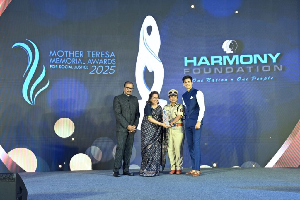 Cuatro personas en el escenario durante los Premios Conmemorativos Madre Teresa para la Justicia Social 2025, otorgados por la Fundación Harmony. Una mujer uniformada sostiene un premio que celebra el poder de la narración, con los logotipos del evento desplegados en una gran pantalla detrás de ella.