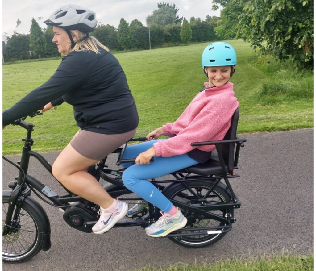 Two people ride a bicycle together on a paved path. The adult in front pedals while a smiling child in a blue helmet and pink jacket sits on the rear seat. Green grass and trees are in the background.