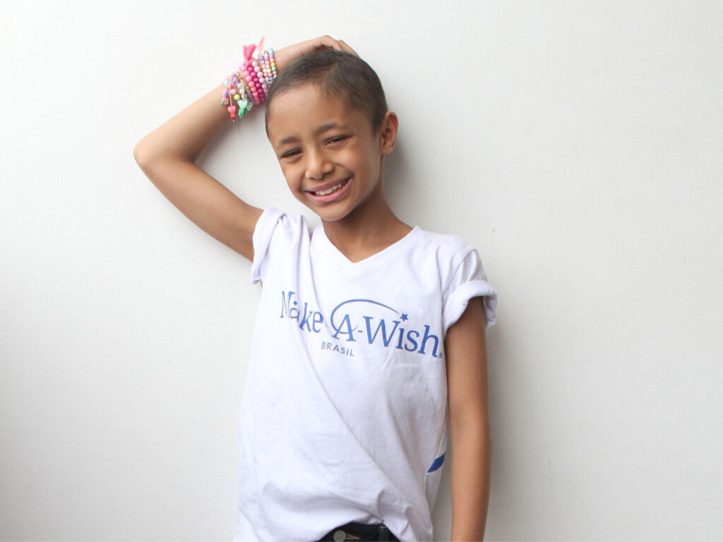 A smiling child with short hair wears a white Make-A-Wish Brasil T-shirt and colorful beaded bracelets, standing against a plain white wall with one hand resting on their head—inviting you to sign to receive the newsletter.