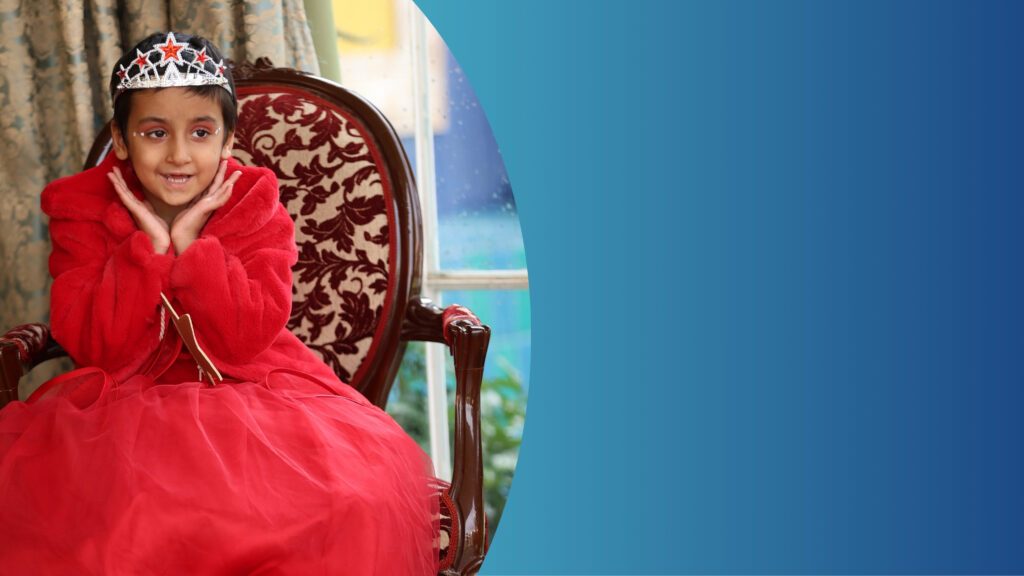 Ein junges Mädchen in einem roten Kleid und mit silbernem Diadem sitzt lächelnd auf einem reich verzierten Stuhl und stützt das Kinn in die Hände. Der Hintergrund ist ein blauer Farbverlauf; links sind ein Fenster und ein Vorhang zu sehen.