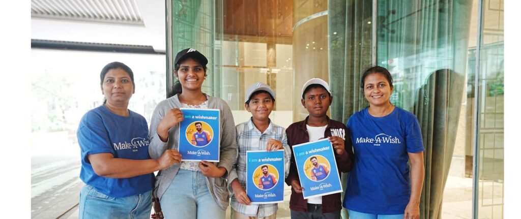 Five people stand and smile at the camera. Two wear blue Make-A-Wish shirts, while three children hold signs featuring a cricketer and the text I am a wishmaker. They appear to be participating in a Make-A-Wish event.