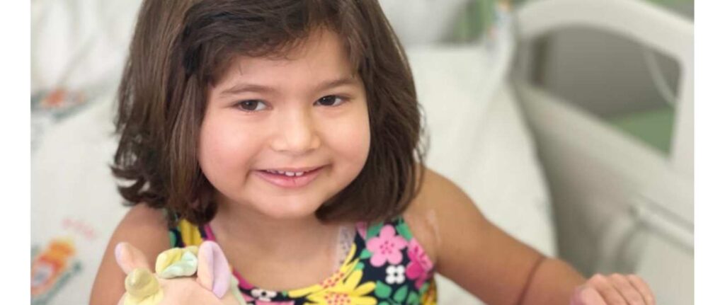 A young girl with brown hair smiles whilst sitting on a hospital bed. She is wearing a colourful floral dress and holding a bandaged hand shaped into a peace sign. The background shows medical equipment and white bedding.