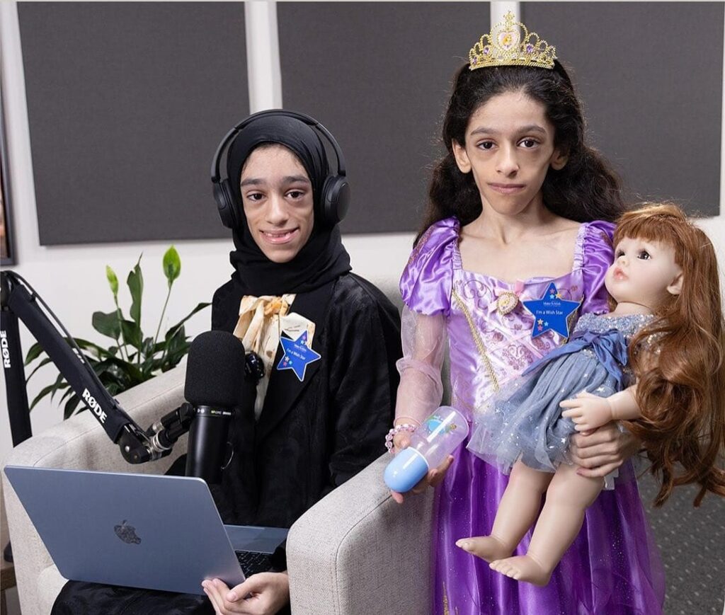 Two young girls sit indoors; one wears a hijab with headphones and sits at a laptop and microphone, while the other wears a princess dress and crown, holding a doll and a toy. Both have name badges and serious expressions.