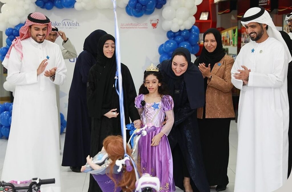 A young girl in a princess dress, embodying Layan’s wish to play all the time, stands surrounded by smiling adults in traditional Middle Eastern clothing, with balloons and a decorated bicycle in the foreground, at a celebration event.