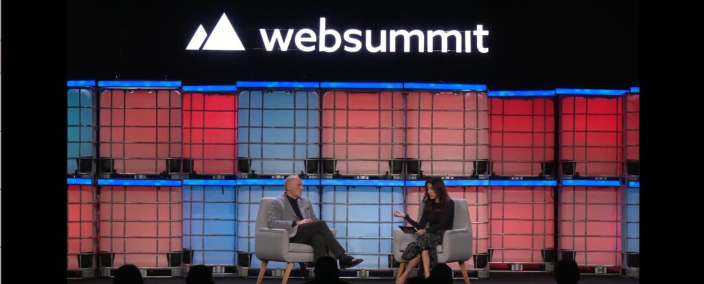 Two people sit and talk on stage at Web Summit, with a backdrop of stacked blue and red crates and the Web Summit logo displayed above them, as they discuss Make‑A‑Wish International being recognised with the Mother Teresa Memorial Award for Social Justice.