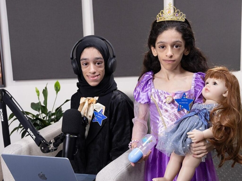Two children sit indoors; one wears a headset and suit at a microphone, and the other, dreaming of Dimitra's wish to have a new room, wears a purple princess dress and crown, holding a doll and bottle. Both have blue star name tags.