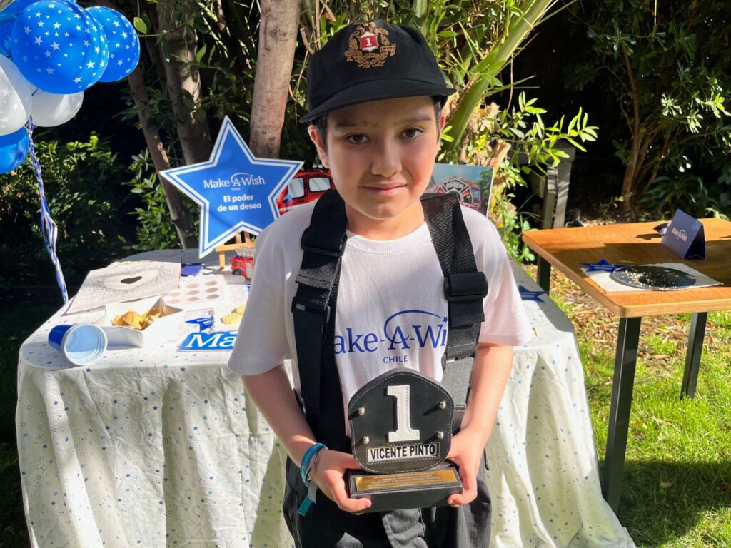 A young boy wearing a black cap and white Make-A-Wish t-shirt holds a “1” trophy with the name “Vicente Pinto,” celebrating Vicente's wish to be a firefighter. He stands by a decorated table with balloons and greenery in the background.