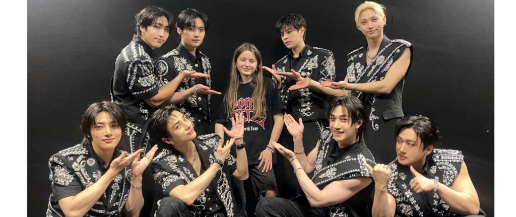 Nine men in matching black, ornate outfits pose on either side of a smiling young woman in casual clothes, all making playful hand gestures towards her against a plain dark background.