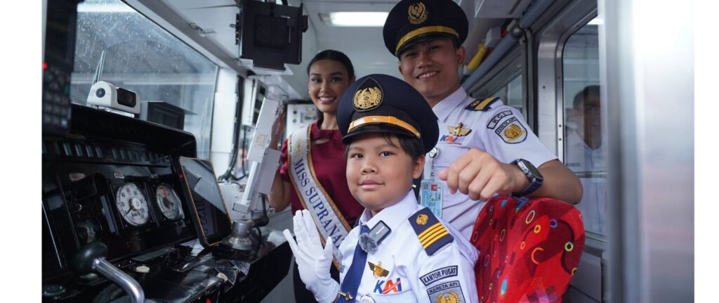 A woman wearing a sash that reads Miss Supranational poses with a smiling man and a child, both in pilot uniforms, inside what appears to be the cab of a vehicle or train.