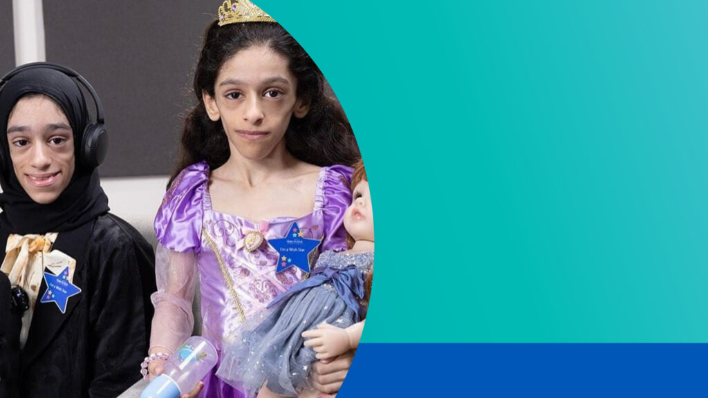 Two children in fancy dress stand side by side. One wears a black outfit with a headscarf and headphones. The other wears a purple princess dress, a tiara, and holds a doll. Both have blue star-shaped badges.