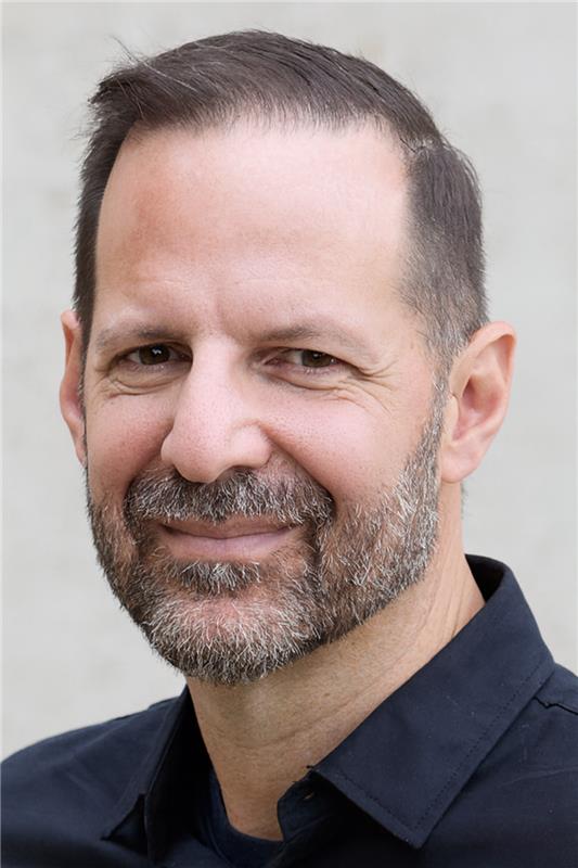 A middle-aged man with short dark hair and a salt-and-pepper beard is smiling slightly. He is wearing a dark collared shirt and is posed against a plain, light-coloured background.