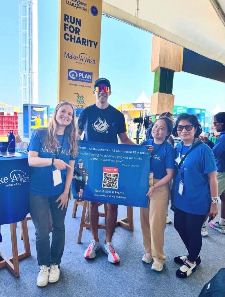 Four people in blue Make-A-Wish shirts stand indoors at a charity event. One man in the centre wears sunglasses and holds a banner about running marathons for charity. Smiling faces and event stalls are visible in the background.