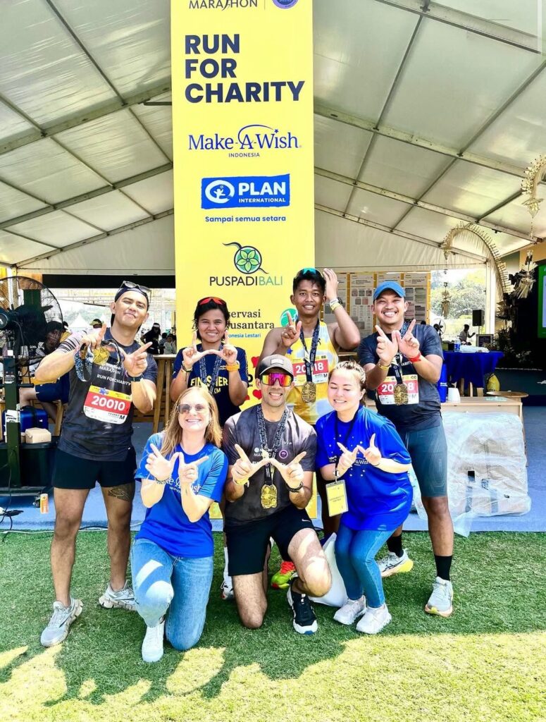 Seven smiling people pose together in front of a yellow “Run for Charity” banner under a marquee; four wear numbered race bibs and medals, while three kneel in blue shirts, all making celebratory hand gestures.