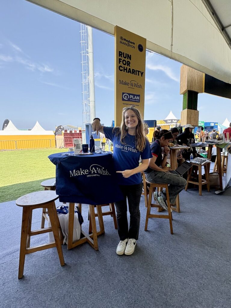 A woman in a blue Make-A-Wish T-shirt stands at a table with Make-A-Wish merchandise, smiling and giving a thumbs up. Behind her is a Run for Charity sign and other people sitting at tables under a marquee.