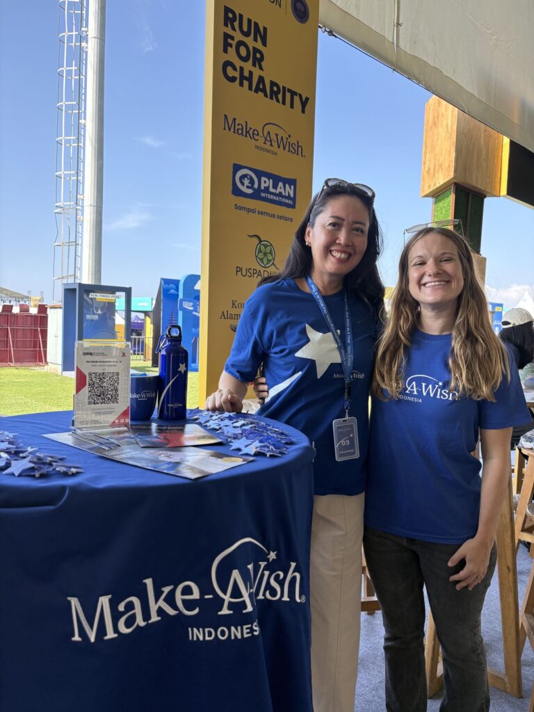 Two women wearing blue Make-A-Wish shirts smile at a stall for a charity run event. The table displays promotional items and a QR code. A yellow event sign and outdoor setting are visible in the background.