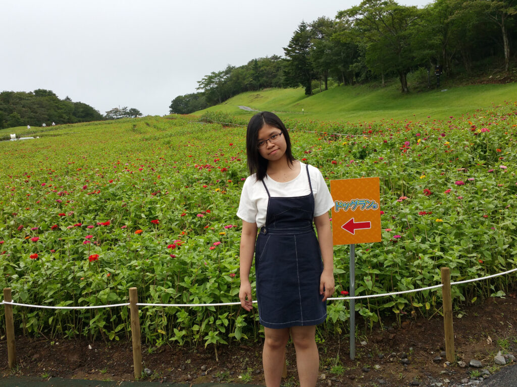 A young woman stands in front of a field of flowering plants with a small orange sign featuring a red arrow. Trees and green hills are in the background under a cloudy sky.
