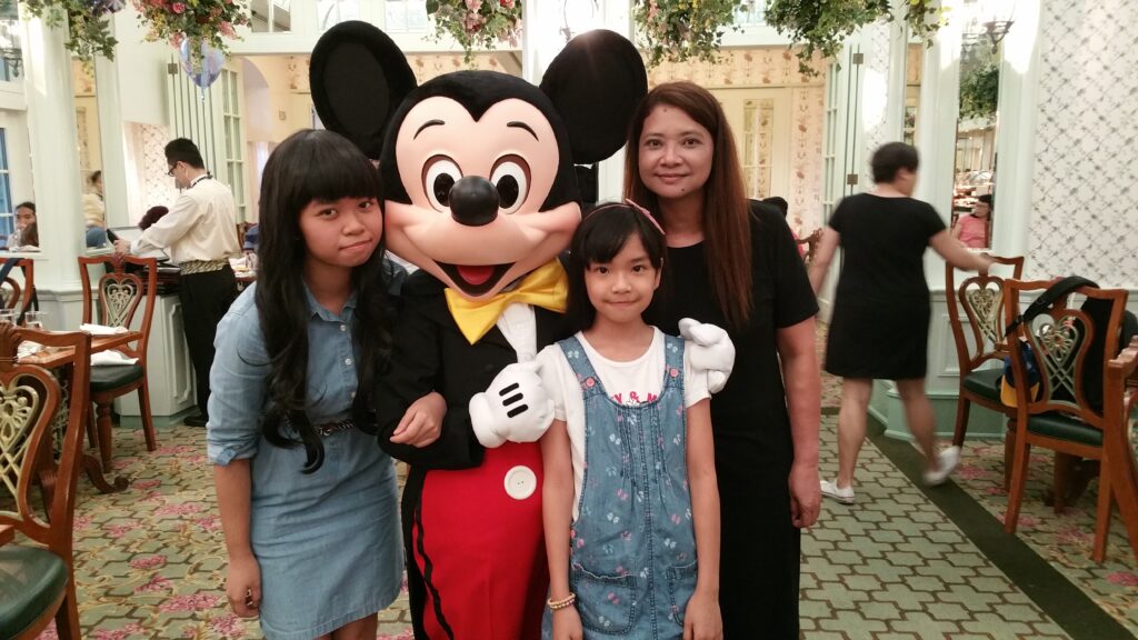 Three children and an adult woman smiling and posing with a person in a Mickey Mouse costume inside a decorated restaurant with wooden chairs and hanging plants.