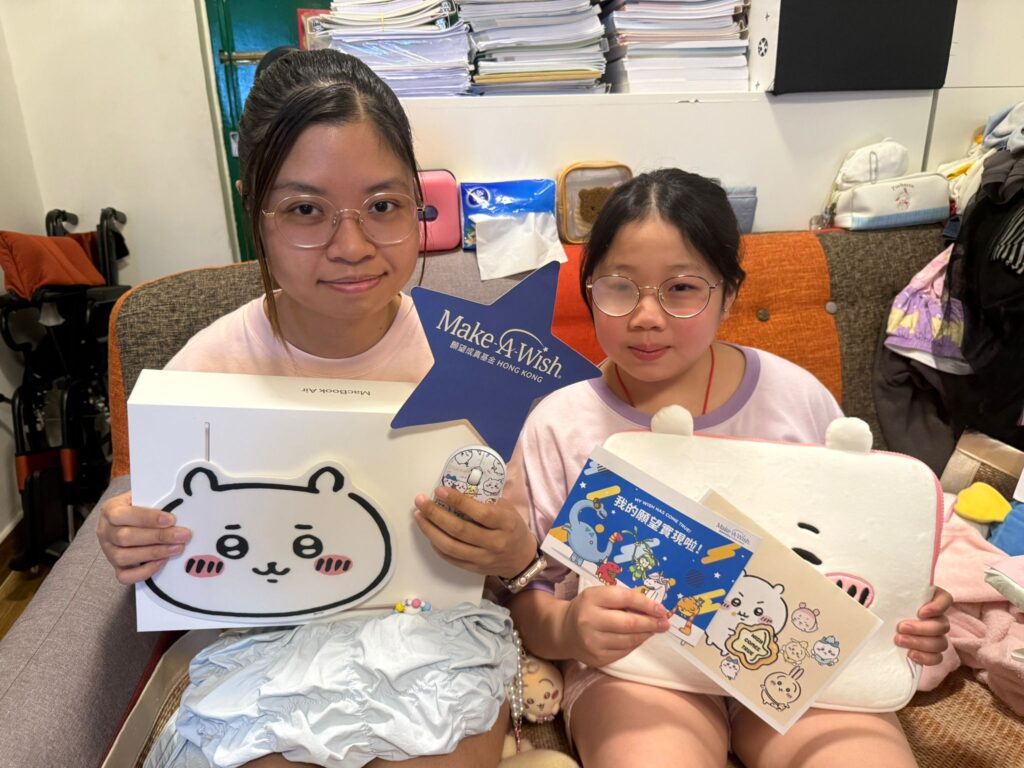 Two girls sitting on a settee hold Make-A-Wish-themed gifts, including a bear-face box, a card with stickers, and a blue star with Make-A-Wish written on it. Piles of books are visible behind them.