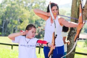 An adult and a child practise archery outdoors. The child, in a Make-A-Wish T-shirt, uses a bow with support from the adult—perhaps sharing their own story as a Wish Mum about getting involved with Make-A-Wish Brazil amid trees and greenery.