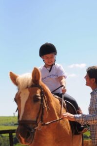 A smiling child wearing a helmet and a Make-A-Wish T-shirt rides a light brown horse, while an adult—perhaps a Wish Mum—holds the reins beside them on a sunny day. Blue sky and greenery are visible in the background.