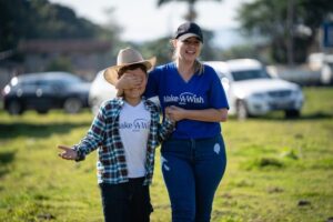 A smiling woman in a Make-A-Wish shirt, a Wish Mum, walks with a child in a cowboy hat and checked shirt. The child covers their eyes, outdoors on a sunny day with cars and trees behind them. Could you share your own story as a Wish Mum?.