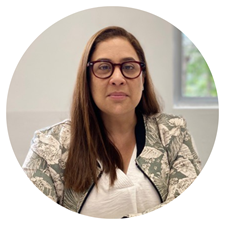 Maria Gracia’s journey with Make-A-Wish began not in an office; here, she sits indoors in front of a window, her long brown hair and glasses complementing a patterned blazer and white top, as she looks thoughtfully at the camera.