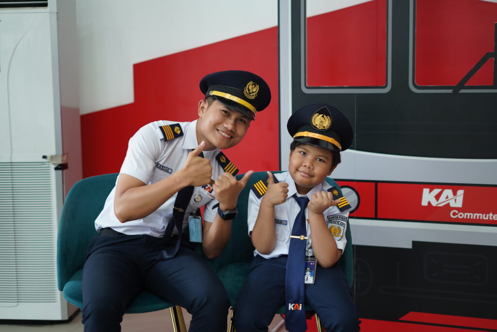A man and a child dressed in matching train conductor uniforms sit on green chairs, smiling and giving thumbs up—capturing Fatah's wish to be a train driver. A red and white wall and part of a train graphic are visible in the background.