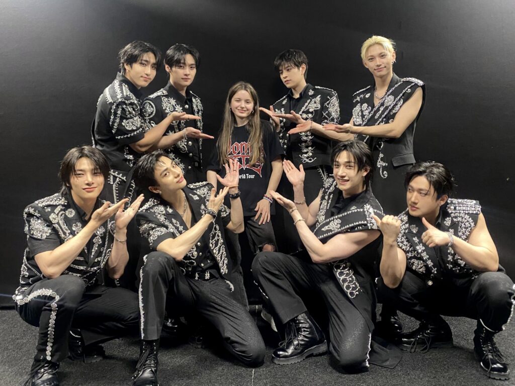 A girl stands smiling in the centre, surrounded by nine men in black, intricately patterned outfits, all pointing towards her and posing enthusiastically—perhaps celebrating Vicente's wish to be a firefighter—against a plain black background.