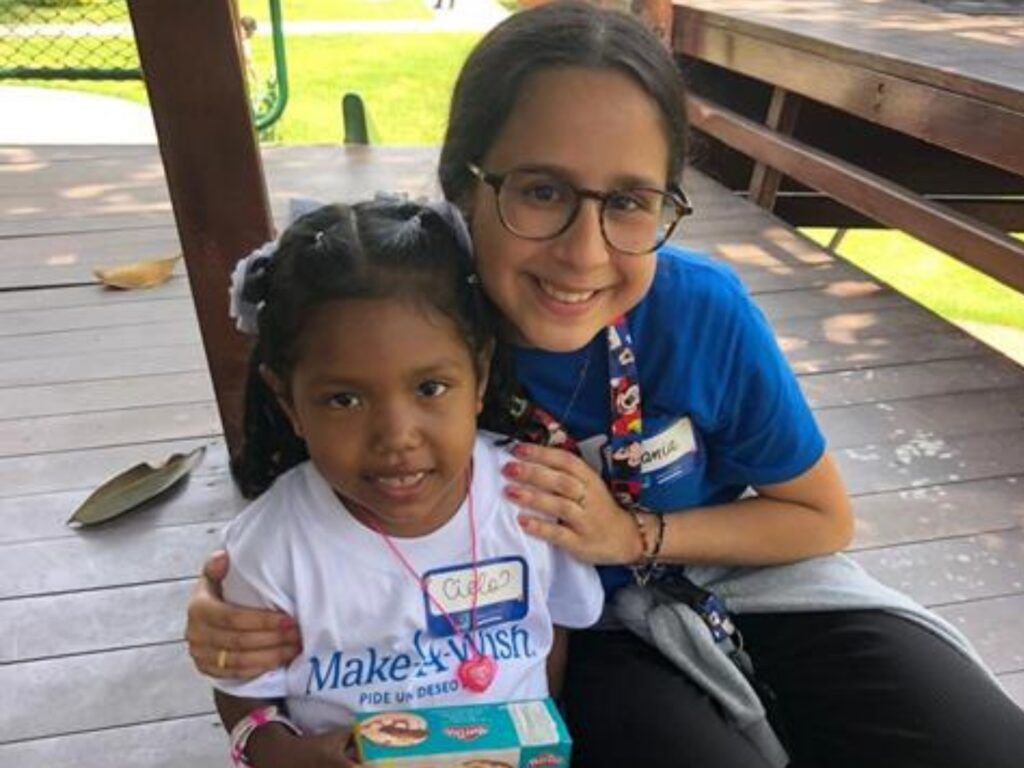 A young girl and an older girl, both smiling, sit together on a wooden veranda. The younger girl wears a Make-A-Wish shirt and holds a toy box, while the older girl in glasses hugs her and wears a blue shirt and lanyard.