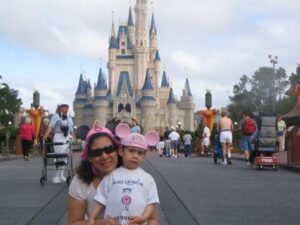 Maria Gracia’s journey with Make-A-Wish began not in an office, but in front of Cinderella Castle, where she and a child wearing Mickey Mouse ear hats smile for a photo as other visitors and pushchairs fill the walkway around them.