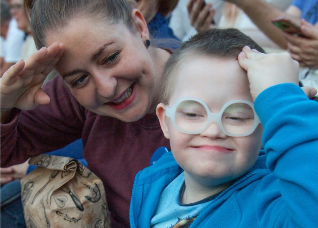 A smiling woman and a young boy with Down’s syndrome wearing glasses both salute playfully, enjoying a happy moment together in a crowd. Kari’s first encounter with Make-A-Wish was when her son was going through leukaemia treatment, an experience that inspired her to become a dedicated volunteer with Make-A-Wish Norway.