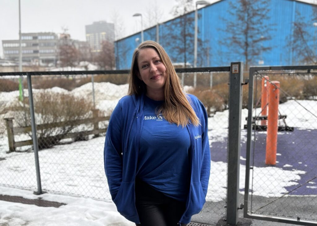 A woman with long brown hair, wearing a blue jacket and T-shirt, stands and smiles outside near a fence. Snow covers the ground, and buildings and trees are visible in the background.
