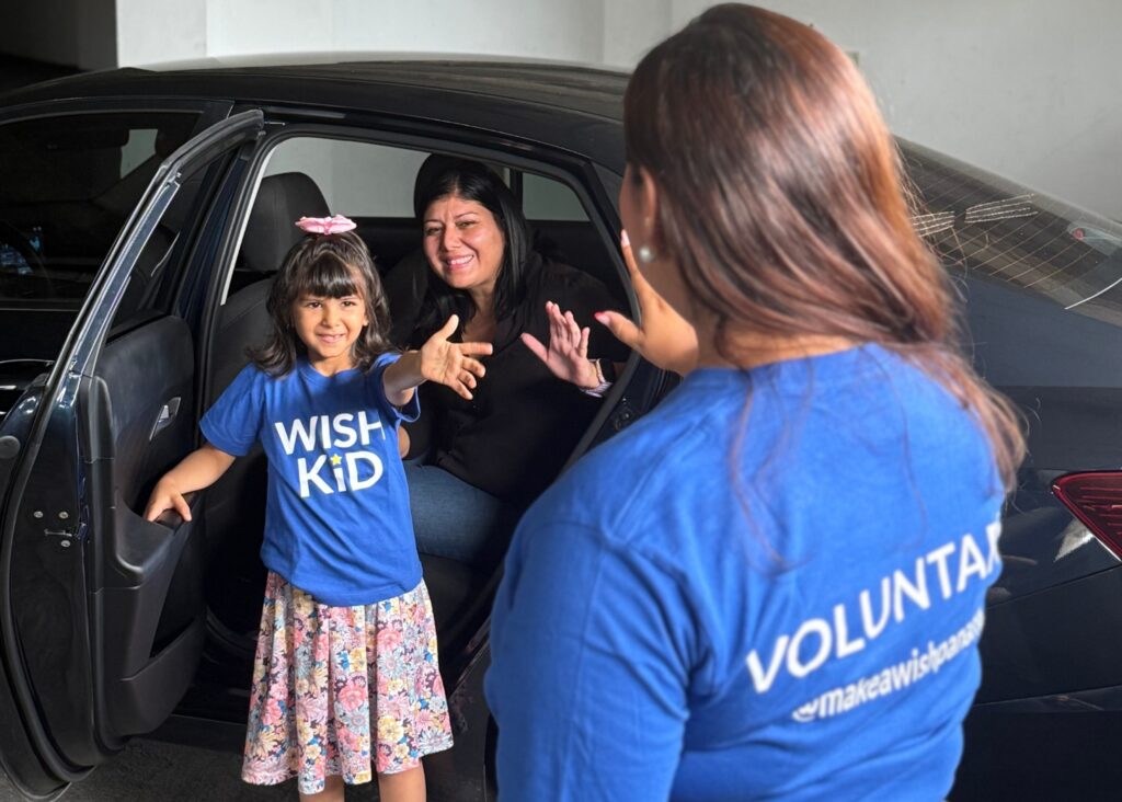 A smiling girl wearing a WISH KID shirt and pink bow steps out of a car, reaching towards a woman in a VOLUNTEER shirt, whilst another woman in the car smiles and waves—one of many inspiring wishmakers’ stories.