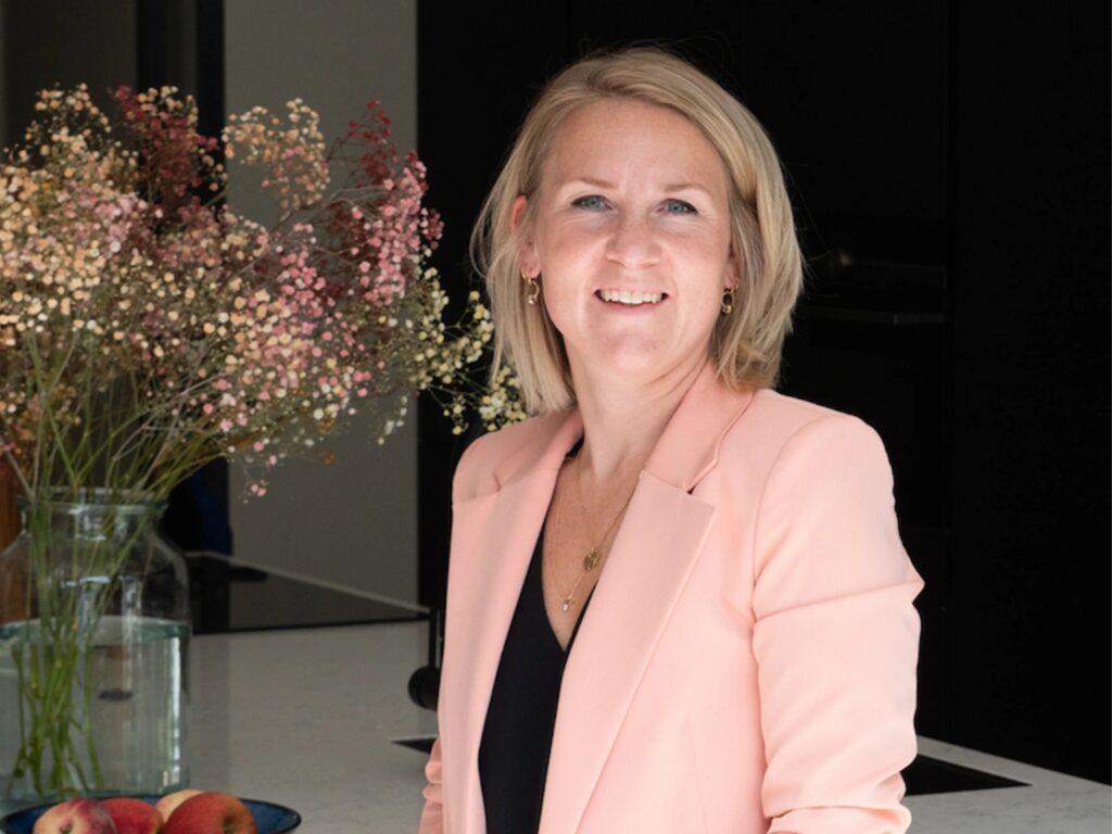 A woman with blonde hair, wearing a light pink blazer and a black top, smiles whilst standing indoors beside a vase of flowers on a white worktop.