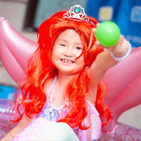 A smiling child in a red wig, tiara, and mermaid costume sits on a pink inflatable, holding up a green ball.