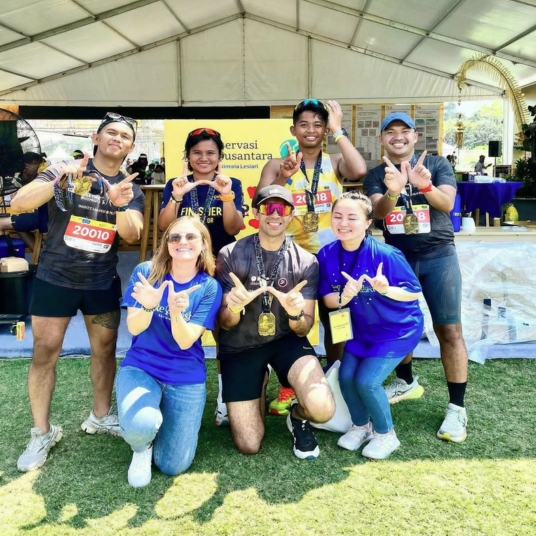 A group of seven smiling people pose outdoors under a marquee, some wearing race numbers and medals. Four are kneeling in front whilst three stand behind, all making “W” hand signs. The atmosphere is cheerful and sunny.