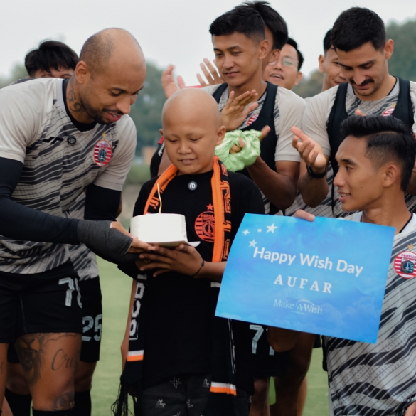 A group of football players surrounds a smiling child holding a small cake. One player holds a blue sign that reads Happy Wish Day AUFAR Make-A-Wish. The group appears joyful and supportive on a sports pitch.