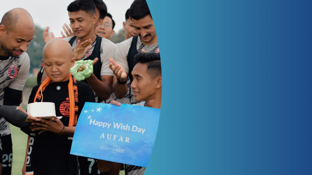 A group of football players gathers around a smiling child holding a small cake. One player holds a sign that reads Happy Wish Day AUFAR Make-A-Wish against a blue gradient background.