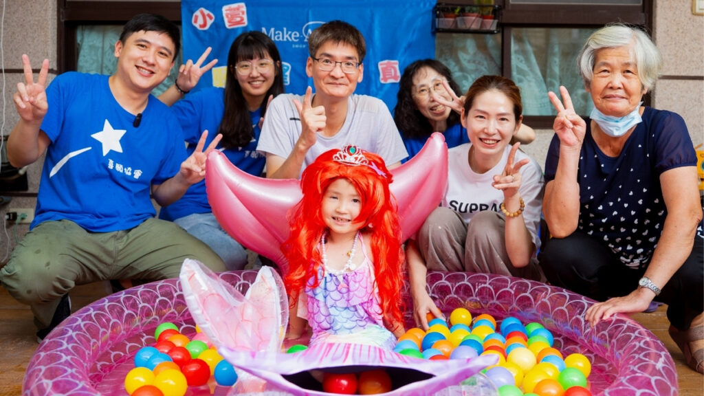 A young child dressed as a mermaid sits in a colourful ball pit, smiling and surrounded by six happy adults, all making peace signs. A blue Make-A-Wish banner is visible in the background.