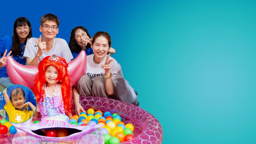 A group of smiling adults and children pose together. A girl dressed as a mermaid sits in a small inflatable paddling pool filled with colourful plastic balls. The background is a blue gradient with empty space on the right.