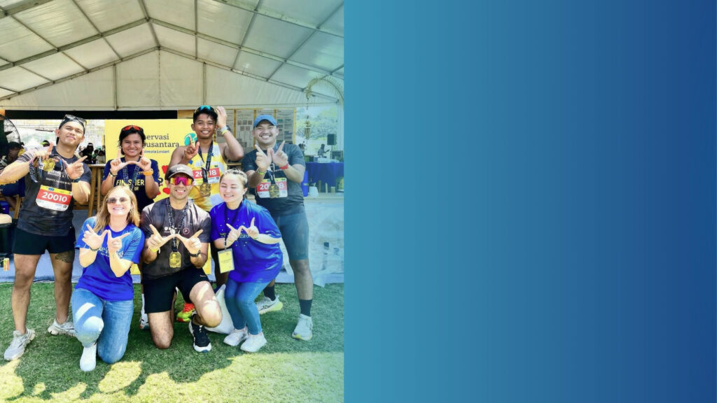 A group of six smiling people, some wearing medals and running numbers, pose under a white marquee outdoors. Four stand, two kneel in front, all making a W hand gesture. Bright sunlight and grass are visible.