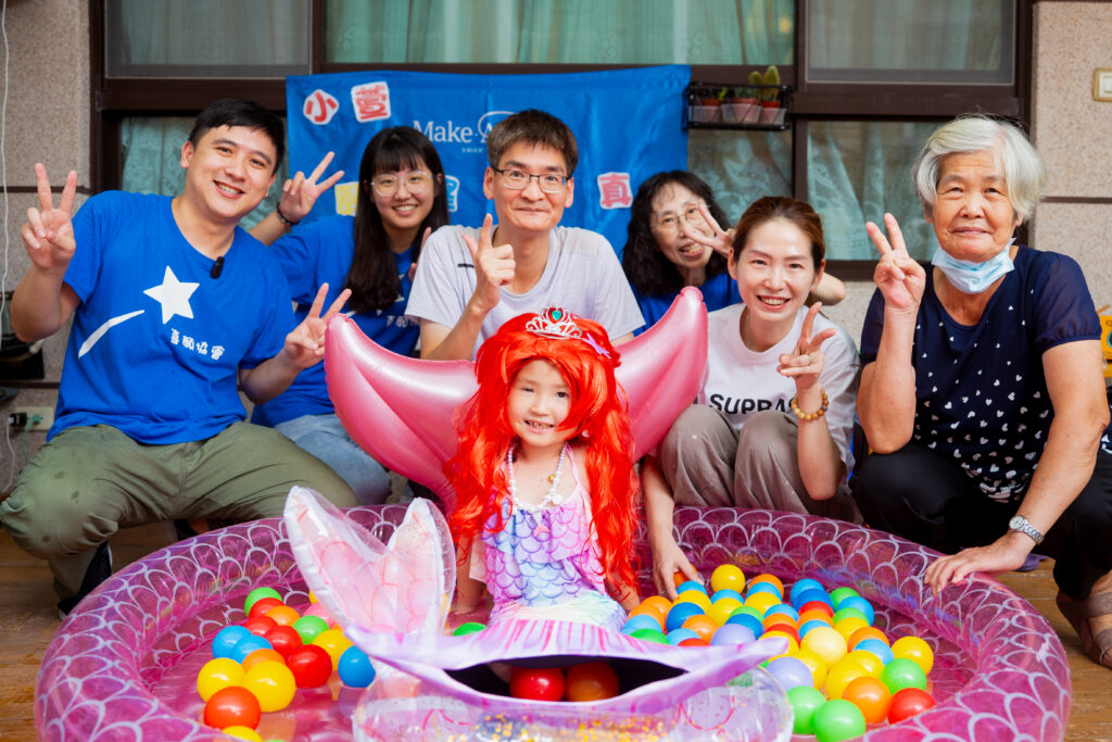A young girl dressed as a mermaid sits in a paddling pool filled with colourful plastic balls, smiling, surrounded by six adults making peace signs and posing happily—celebrating Loraine's wish to meet Stray Kids in a joyful indoor gathering.