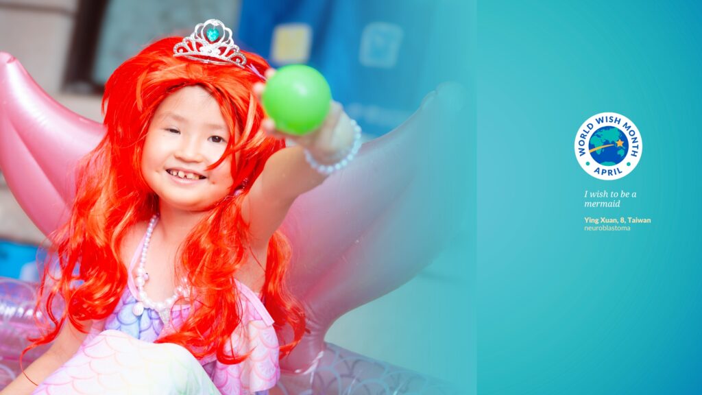 A smiling child with a red mermaid wig and crown, wearing a pearl necklace and mermaid tail, sits in a pink shell chair, holding out a green ball. Text reads: World Wish Month April. I wish to be a mermaid.