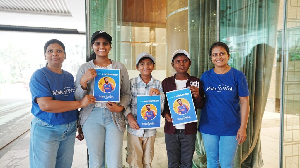 Five people stand together indoors, smiling at the camera; three children in the centre—celebrating Ying Xuan's wish to be a mermaid—hold I am a wishmaker posters, whilst two women in Make-A-Wish shirts stand on either side, supporting the children.