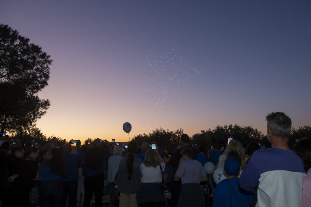 A crowd watches at dusk as a star shape appears in the sky, formed by illuminated drones. Many people are taking photos or videos with their mobiles. Trees are silhouetted against the sunset.