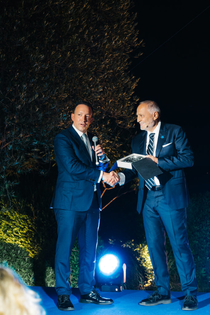 Two men in suits stand on a blue-lit outdoor stage at night. One man holds a microphone whilst shaking hands with the other, who holds a folder. A tree and greenery are visible in the background.