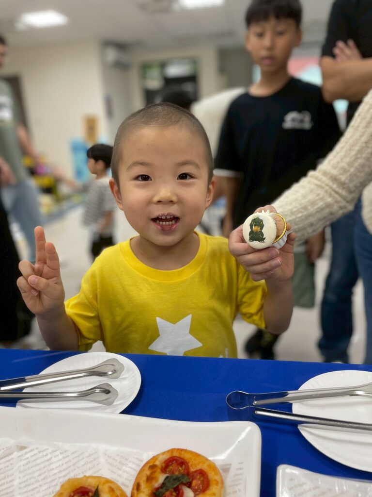 A young child in a yellow shirt with a white star smiles and makes a peace sign while an adult holds a decorated biscuit near him, joyfully sharing Adarsh's wish to meet Rohit Sharma. Plates of pastries are on the table, people stand in the background.