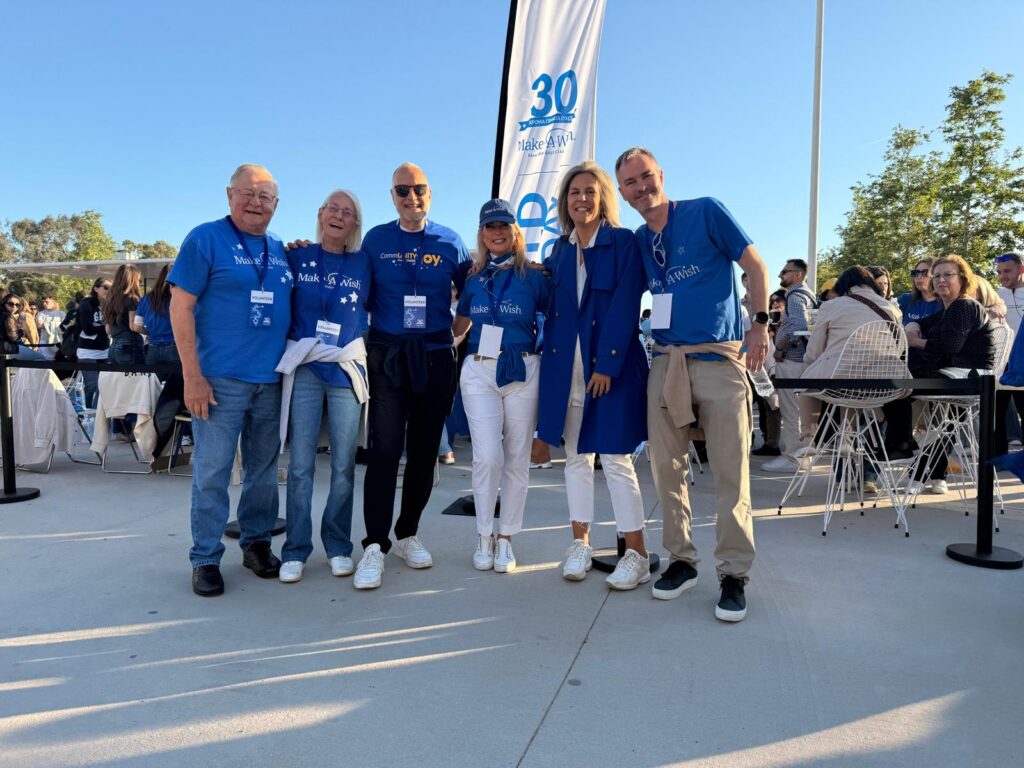 Six people wearing blue and white clothes pose and smile together outdoors at an event. A tall banner with 30 and Make-A-Wish is visible in the background, and others are gathered round tables behind them.