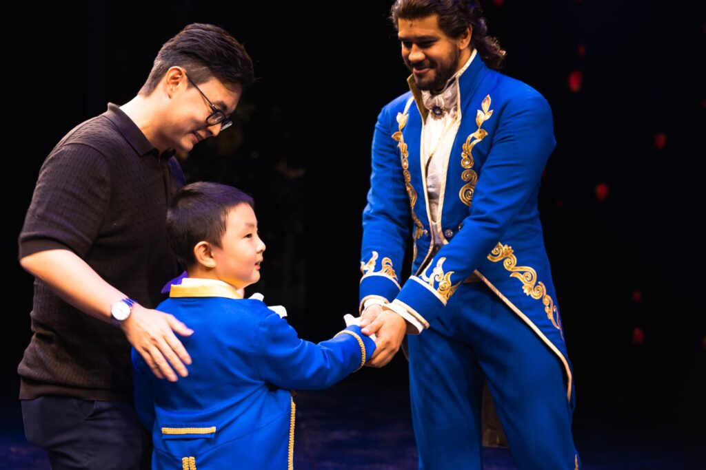 A man and a child, both smiling, shake hands with a performer in an ornate blue and gold costume on stage. The child also wears a blue costume and looks excited. The background is dark with hints of red lights.