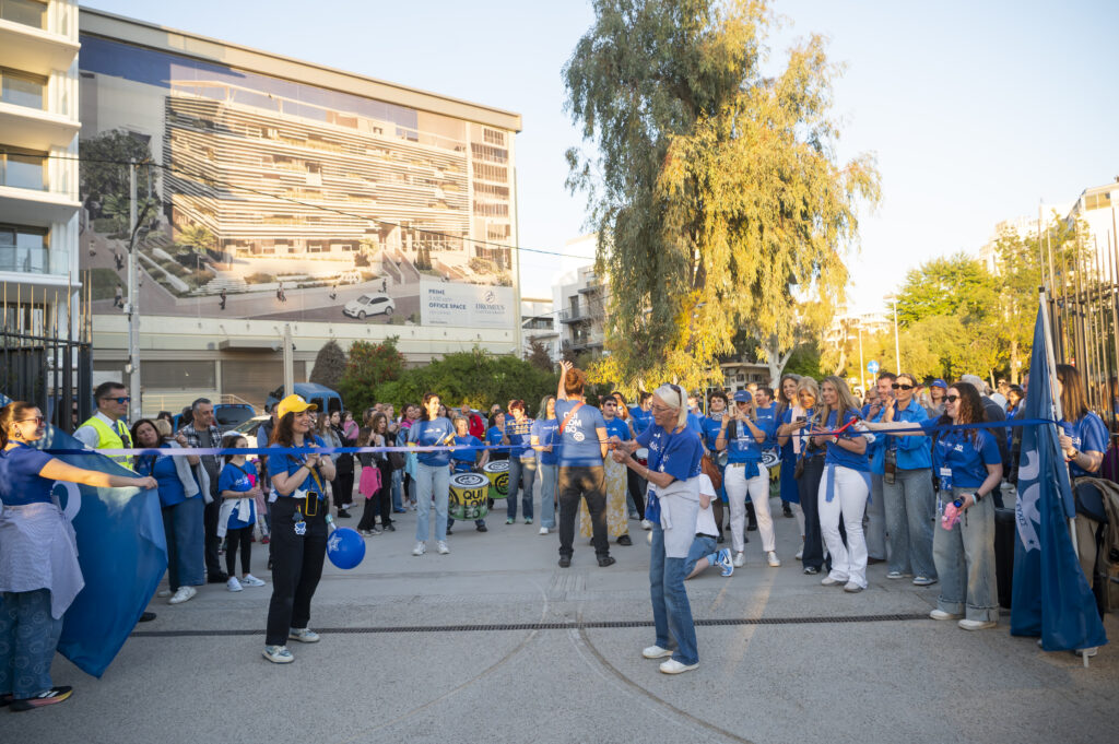 A group of people in blue shirts gather outdoors for a ribbon-cutting ceremony, surrounded by balloons and cheering participants. A large building and mural are visible in the background.